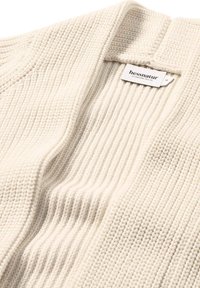 Strickjacke in Beige mit einem strukturierten Rippmuster. Enthält ein Etikett mit "hessnatur"-Markierung nahe dem Ausschnitt.