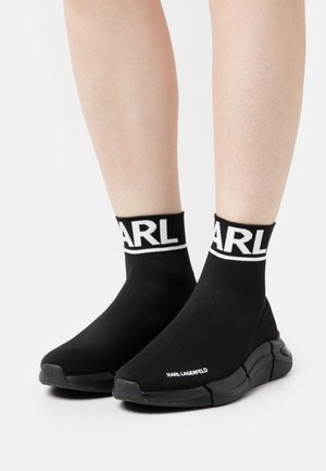 Baskets style chaussette noires avec une tige en maille, une bande blanche avec logo, et une semelle en caoutchouc épurée. Elles présentent un bout rond et un design minimaliste.