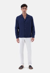 Camicia blu navy con colletto a camicia, maniche lunghe e tessuto texturizzato, abbinata a pantaloni bianchi e sandali neri.