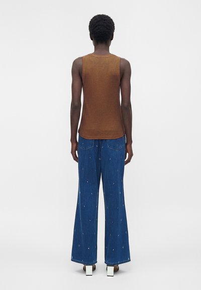 Άτομο με κοντά σγουρά μαλλιά που φορά καφέ αμάνικο τοπ, φαρδιά μπλε τζιν με μικρά λευκά σχέδια και λευκά παπούτσια με κλειστό τακούνι, φαίνεται από πίσω.