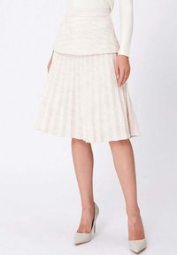 Femme portant une jupe plissée rose pâle arrivant au genou avec une taille cintrée et des chaussures à talons hauts blanches, se tenant devant un fond uni.