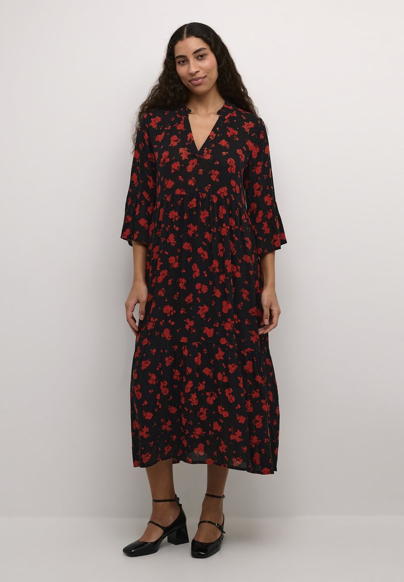Robe midi noire à motifs floraux avec des fleurs rouges, décolleté en V, manches trois-quarts, et ourlet à volants superposés, associée à des talons noirs.