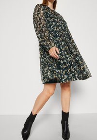 Květnová šaty v tmavě zelené barvě, s dlouhými rukávy a plisovaným designem. Černé kotníkové boty doplňují outfit, s detailem výstřihu ve tvaru klíčové dírky.