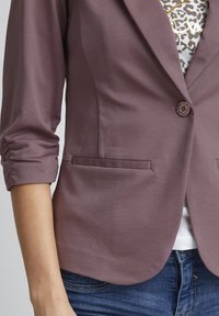 Fialový blazer s jedným gombíkom, opätovne ohnutými rukávmi a dvoma prednými vreckami. Hladká látka s jemným leskom a štruktúrovaným strihom.