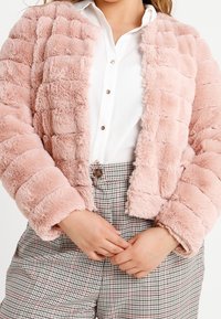 Lyserød faux fur-jakke med et quiltet design, lange ærmer og en cropped pasform, parret med en hvid skjorte og ternede bukser.