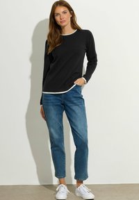 Schwarz langärmliges Hemd mit weißer Kante, kombiniert mit blauen Jeans und weißen Turnschuhen. Das Hemd hat einen runden Ausschnitt und eine lockere Passform.