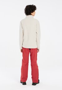 Chaqueta de forro polar en beige claro con un alto cuello y mangas largas, combinada con pantalones impermeables rojos, con un corte holgado y cremalleras.