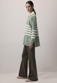 Mintgroene oversized trui met witte horizontale strepen, gecombineerd met bruine wijde broek en zachtroze schoenen. Gele kraagdetails zijn zichtbaar.