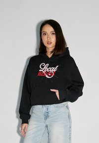 Czarna skrócona bluza z kapturem z różowym napisem "Lokalni Bohaterowie", z przednią kieszenią i luźnym krojem, w połączeniu z jasnoniebieskimi dżinsami.