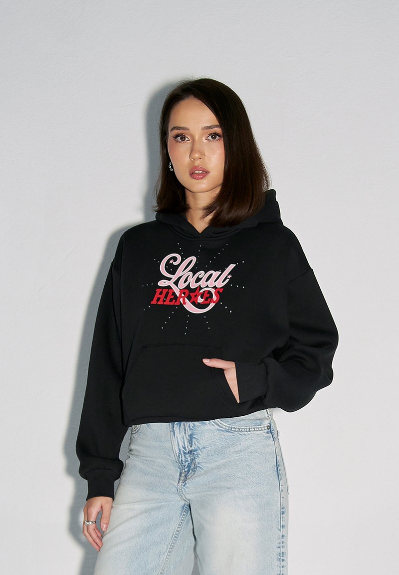 Czarna skrócona bluza z kapturem z różowym napisem "Lokalni Bohaterowie", z przednią kieszenią i luźnym krojem, w połączeniu z jasnoniebieskimi dżinsami.