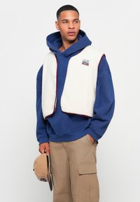 Fleece vest in cream with maroon trim over a blue hoodie. Beige cargo pants and holding a tan cap. Casual layered outfit.