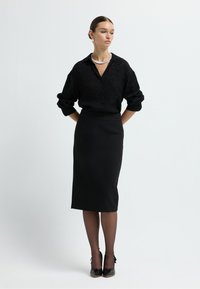 Schwarzes Hemd mit dezentem Muster, in einen figurbetonten schwarzen Bleistiftrock gesteckt, kombiniert mit schwarzen hohen Schuhen und transparenten Strumpfhosen.