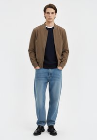 Giacca bomber marrone sopra un maglione blu navy, abbinata a jeans azzurri a gamba dritta e scarpe nere. Design semplice con colori a contrasto.