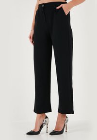 Black, tailored trousers with a high waist, featuring front pockets and a smooth texture. Paired with black heels that have a patterned accent.