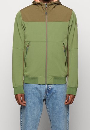 Homme portant une veste à capuche zippée verte et marron avec des poches latérales zippées et un jean bleu clair, se tenant devant un fond uni.