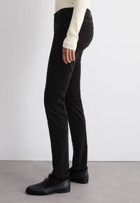 Pantalons en velours côtelé noir avec une coupe ajustée, présentant une taille haute, des poches latérales et une texture subtile. Associés à des chaussures noires à enfiler.