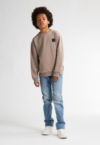 Felpa beige con una vestibilità rilassata, polsini a coste e una piccola etichetta nera sul petto, abbinata a jeans azzurri e scarpe da ginnastica bianche.