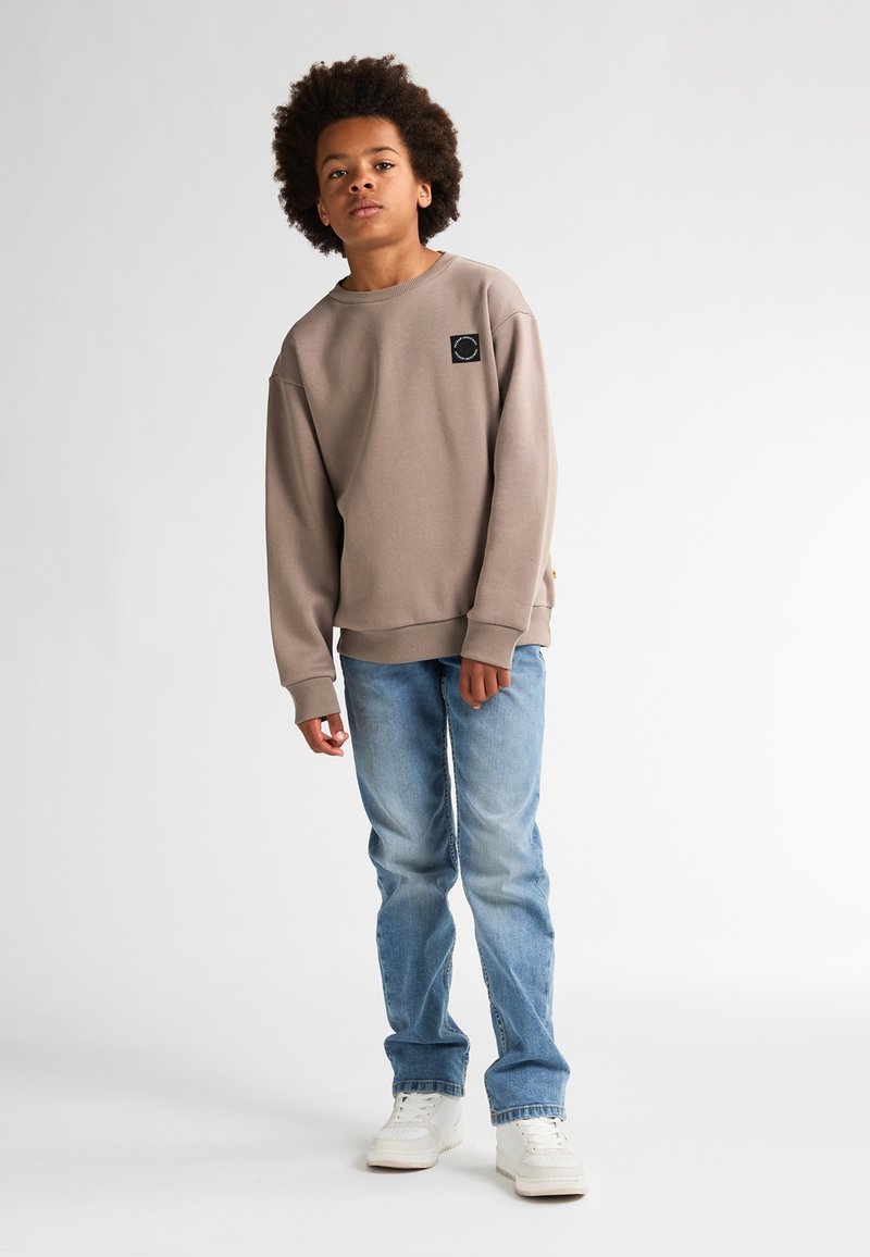Felpa beige con una vestibilità rilassata, polsini a coste e una piccola etichetta nera sul petto, abbinata a jeans azzurri e scarpe da ginnastica bianche.