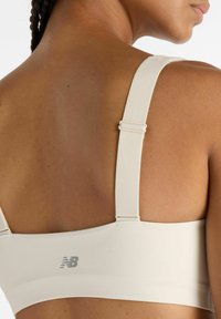 Top deportivo beige claro con tirantes anchos, ajuste con herrajes regulables y textura suave. Presenta un logo gris sutil en la banda trasera.