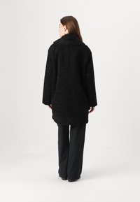 Cappotto nero, texture con un alto colletto e spalle larghe, che si estende oltre i fianchi. Indossato sopra pantaloni neri, che mostrano una vestibilità rilassata.