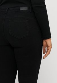 Person med svarte tettsittende jeans og svart langermet topp, viser baklommen og høyrehånden hengende avslappet ved siden.