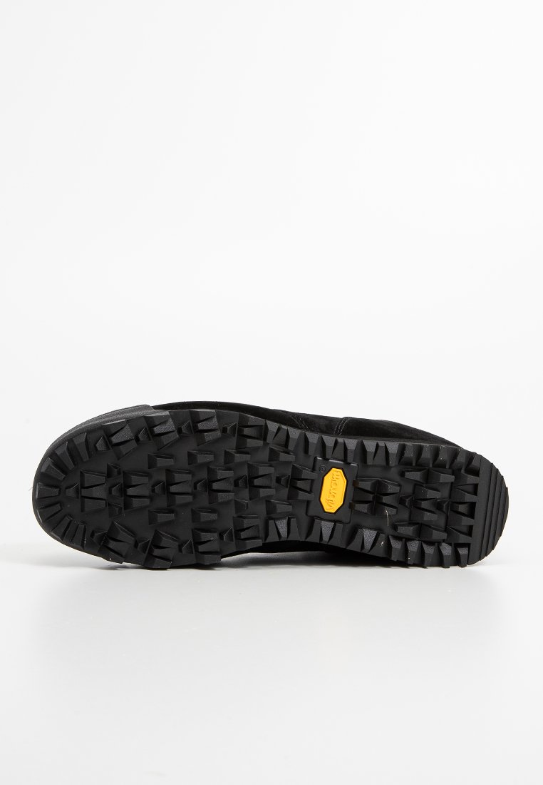 La suola di una scarpa da esterno nera presenta un motivo in gomma ruvido e texturizzato con profondi tasselli per la trazione, messo in risalto da un logo Vibram giallo.