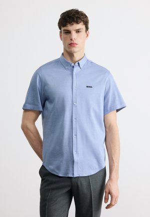 Mladi moški v svetlo modri srajci s kratkimi rokavi z logotipom »BOSS« na prsih in temno sivih hlačah, postavljen pred belo ozadje.