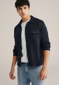 Mörkblå stickad kofta med knappraden fram, två bröstfickor, ribbade muddar och en mjuk textur, lagd över en vit t-shirt.