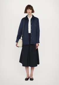 Marineblå skjortejakke over en hvid skjorte, parret med en mørk nederdel. Sorte flade sko og en cremefarvet taske fuldender outfittet. Glat tekstur.