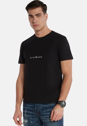 T-shirt in cotone nero con collo rotondo e maniche corte, caratterizzato da un logo bianco "Richmond" al centro. Indossato con jeans blu strappati.