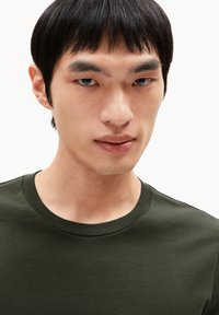Dark green cotton t-shirt with a round neckline. Features short sleeves and a smooth texture, displayed on a model's upper body.