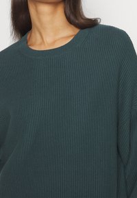 Maglione turchese a costine con un ampio scollo a giro e una vestibilità comoda. La texture è morbida e lavorata a maglia, con maniche lunghe.
