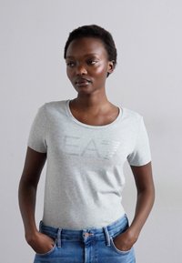 Tricou din bumbac gri, cu mâneci scurte, guler rotund și un logo metalic "EA7" imprimat pe față. Purtați cu blugi denim albaștri.
