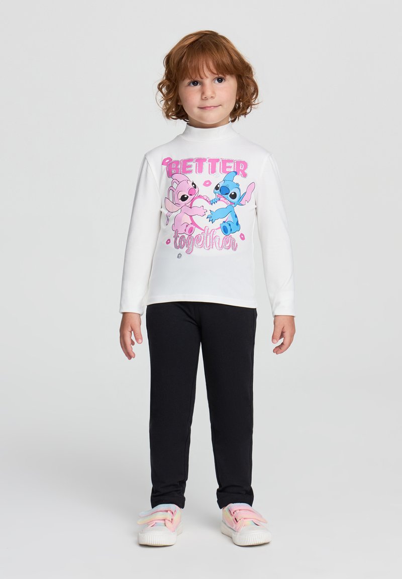 Camicia bianca a maniche lunghe con una grafica colorata che presenta personaggi dei cartoni animati, abbinata a leggings neri e scarpe dai colori pastello.