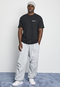 Svart bomull T-skjorte med logo, grå nylon cargo bukser og hvite joggesko. Modell iført grå strikket lue, står på en myk blå overflate.