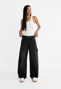 Pantaloni in denim neri a gamba larga con vita alta, chiusura frontale a bottoni e tasche laterali, abbinati a una maglietta aderente bianca e sneaker bianche.