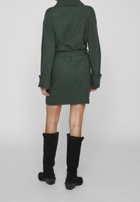 Femme portant une robe en maille vert foncé ceinturée et des bottes noires montantes jusqu'aux genoux, debout devant un fond gris clair uni.