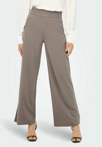 Bredde benbukser i myk, taupe stoff. Elastisk midjebånd med sidelommer og en glatt tekstur, kombinert med åpne svarte hæler.