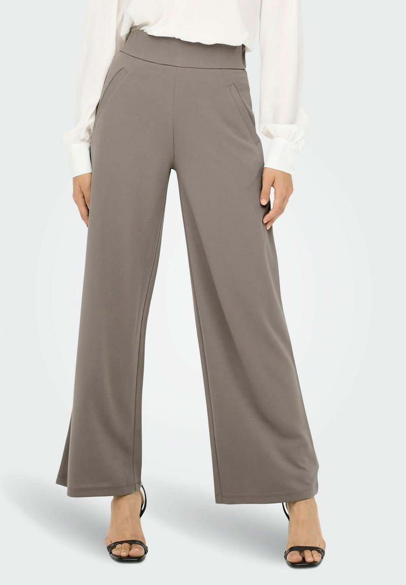 Bredde benbukser i myk, taupe stoff. Elastisk midjebånd med sidelommer og en glatt tekstur, kombinert med åpne svarte hæler.