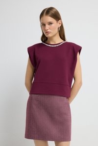 Kvinde med glat lysebrunt hår iført en ærmeløs lilla top med rhinstene ved halsen og et mønstret lilla nederdel, som står op ad en hvid væg.