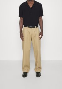 Maglietta nera a maniche corte, pantaloni beige plissettati, cintura nera e scarpe nere. Design semplice con vestibilità comoda e texture liscia.