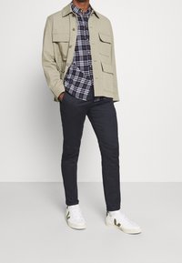 Man wearing beige jacket, navy plaid button-up shirt, dark pants, and white sneakers standing against plain white background.