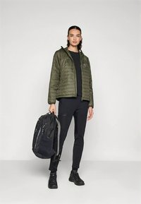 Zöld steppelt kabát kapucnival, fekete felső és nadrág, sötét hátizsákot tart. Fekete cipő texturált talppal. Egyszerű kialakítás és anyagok.