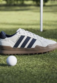 Λευκό και ναυτικό παπούτσι γκολφ δίπλα σε μπάλα γκολφ σε πράσινο γρασίδι κοντά σε σημαία γκολφ.