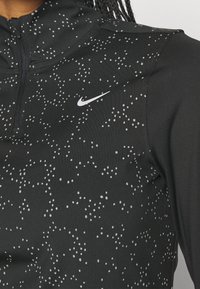 Top sportivo nero con colletto con zip, caratterizzato da un motivo a macchie grigie e un logo Nike bianco sul petto. Tessuto morbido e elasticizzato.