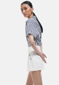 Camisa de manga corta a rayas en azul marino y blanco con cuello, combinada con una falda de mezclilla blanca con botones laterales, modelo de pie con el cabello liso.