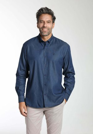 Homme aux cheveux foncés et à la barbe, portant une chemise bleu marine boutonnée et un pantalon beige clair, debout avec une main dans la poche sur un fond blanc.