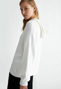 Felpa bianca con cappuccio, dalla texture morbida, vestibilità comoda e polsini a coste. Presenta un cappuccio con cordini e un design minimalista.