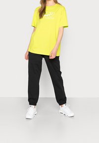 Neon gul t-shirt med en vit tryckt logotyp; parat med svarta avsmalnande joggers. Vita sportskor med synliga luftdämpningdetaljer.