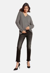 Grauer V-Ausschnitt-Pullover mit langen Ärmeln, kombiniert mit schwarzen, figurbetonten Jeans. Modell trägt schwarze High Heels und steht vor einem neutralen Hintergrund.